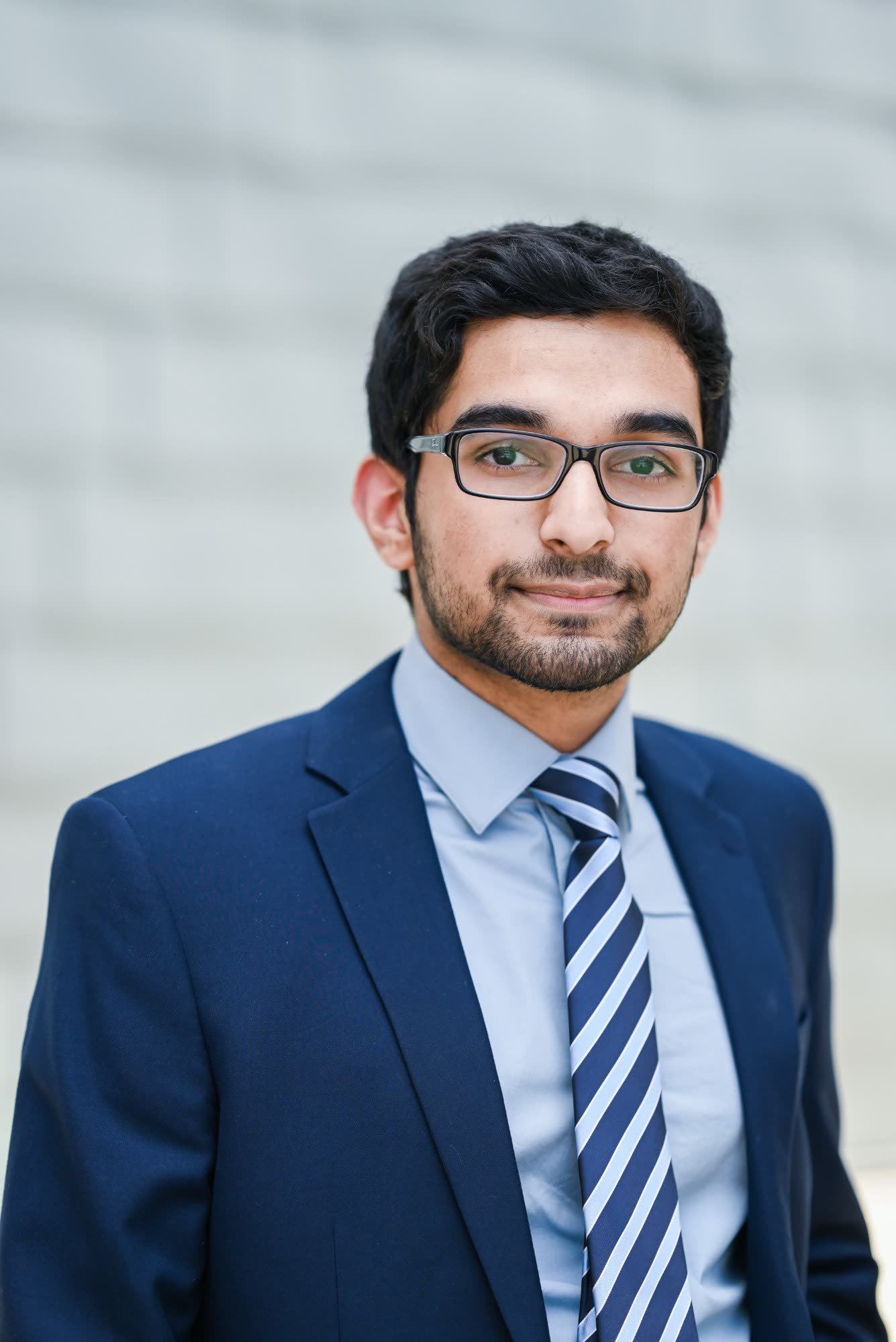 Nabeen Ahmed in a navy suit against a pale brick wall