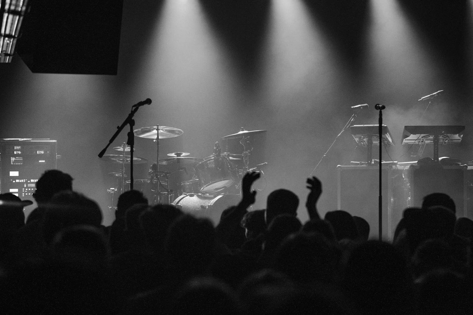 A crowd with hands raised in front of a rock stage, drum kit lit from above